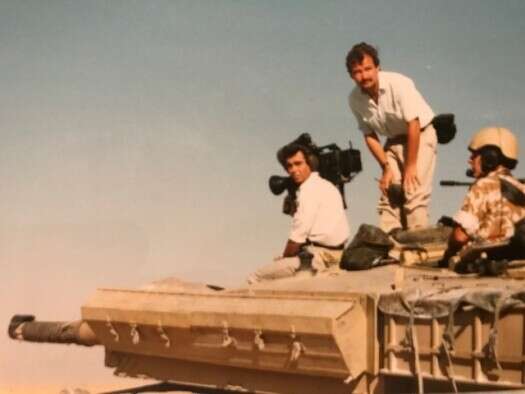 Davies stands atop a tank during the first Gulf War, looking toward camera. A cameraman sits ahead of him, facing toward the distance, next to the tank's main gun. A soldier looking away from them is protruding top half out of the tank hatch.