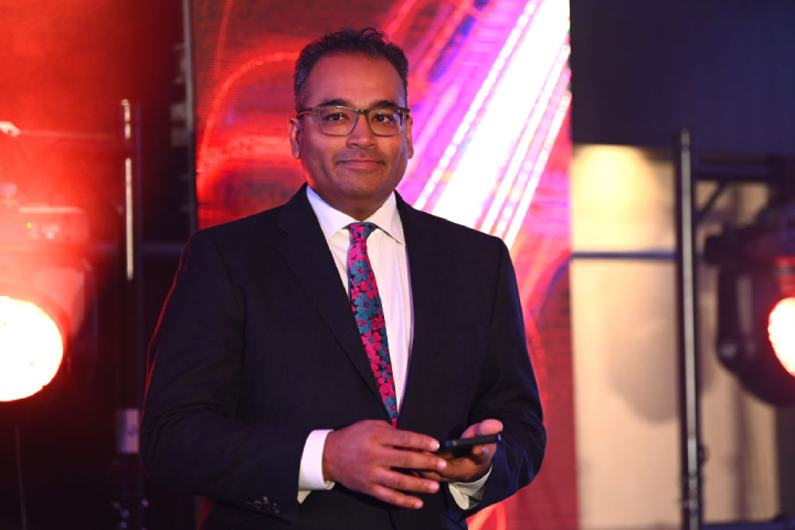 Krishnan Guru-Murthy dressed in a suit and tie smiling at the camera with a bright stage backdrop