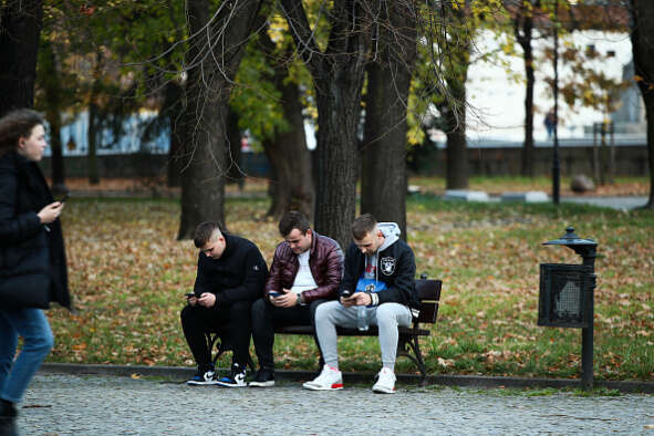 Three young lads sitting on a bench looking at their phones. Now for some keywords for SEO purposes: Gen Z conspiracies Covid News Movement Oliver Wyman