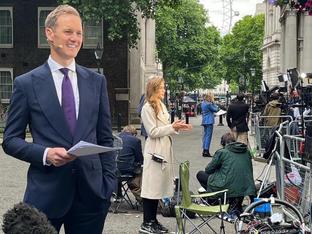 Dan Walker dressed in a suit and holding a stack of paper outside 10 Downing Street, with other journalists in the background
