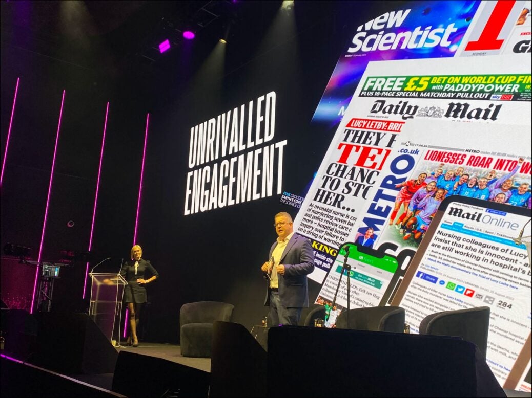 Mail Metro Media chief revenue officer Dominic Williams appears on stage at an advertiser upfront in October 2023. Behind Williams a very large, floor-to-ceiling LED screen is lit up with the words: "UNRIVALLED ENGAGEMENT" and an illustration showing pages from the publications covered by Mail Metro Media.