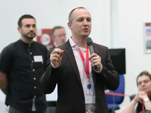 James Scurry standing up at the Media Strong event, wearing a suit and lanyard and holding a microphone