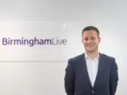 Graeme Brown, the editor of Birmingham Live, is depicted stood smiling in a suit in front of a sign for the website.