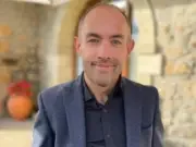 Immediate video boss Paul Doyle smiles at the camera dressed in a shirt and blazer standing in front of a European backdrop of yellow brickwork