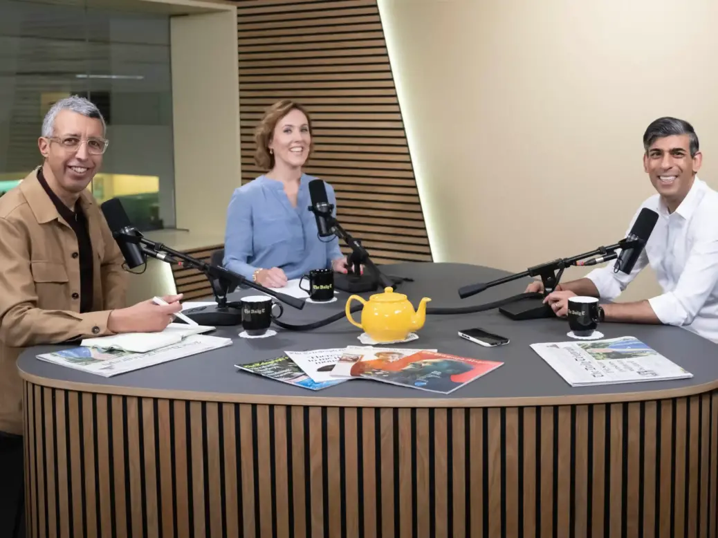 Rishi Sunak visits the studio of The Daily Telegraph's new podcast, The Daily T, to give his first interview of the 2024 general election campaign trail. Kamal Ahmed and Camilla Tominey, the podcast's hosts, sit to the left of a table fitted with microphones while the prime minister sits on the right. Copies of The Telegraph and a yellow teapot are spread over the top of the table.