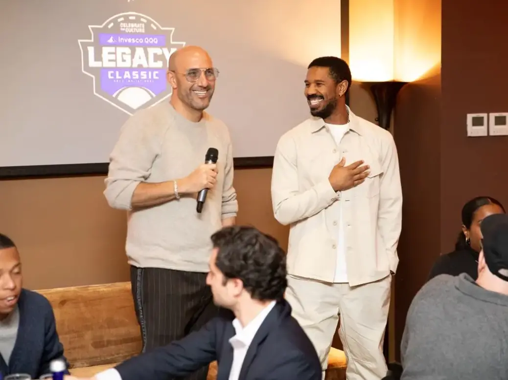 Boardroom co-founder Rich Kleiman stands with a microphone next to actor Michael B. Jordan who holds his hand on his heart and smiles at Kleiman. They're standing in front of the Legacy Classic logo on a projector