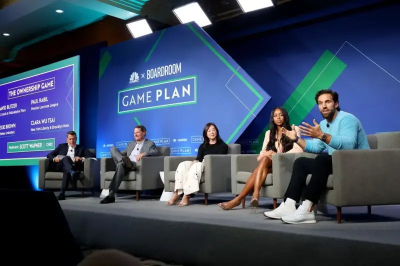 Five people sit on stage in front of a CNBC x Boardroom Game Plan logo - the man on the end is speaking and the rest appear to be listening