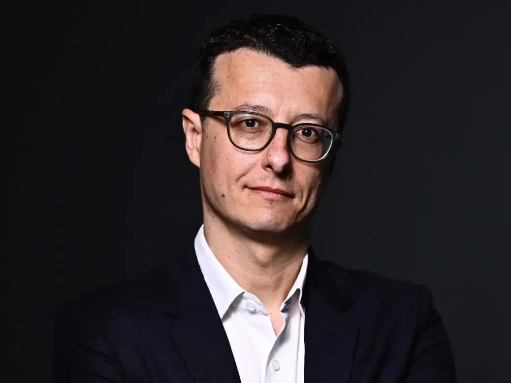 Mehdi Lebouachera, a man wearing glasses and a shirt and blazer facing the camera with crossed arms in front of a black backdrop