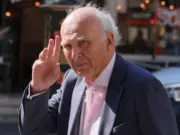 Sir Vince Cable dressed in a suit walking on a street doing a salute gesture at the camera