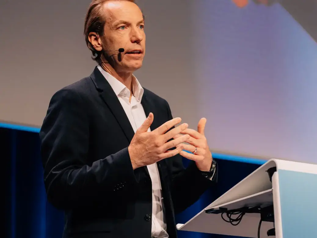 Anders Eriksson speaking on stage standing up gesturing with his hands, wearing a black blazer and white shirt. Blue backdrop, corner of WAN-IFRA branded podium in shot