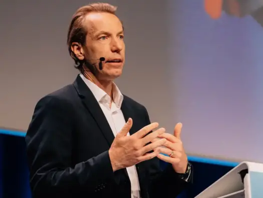 Anders Eriksson speaking on stage standing up gesturing with his hands, wearing a black blazer and white shirt. Blue backdrop, corner of WAN-IFRA branded podium in shot