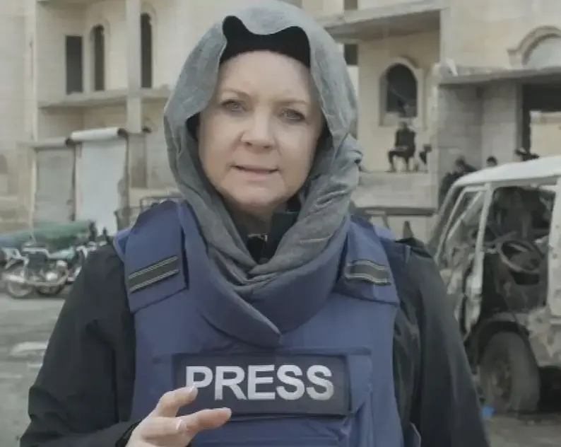Emma Murphy dressed in flak jacket labelled 'press' and head covering, giving piece to camera