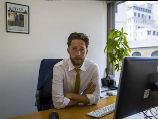 Outgoing City AM editor Andy Silvester leaning on his corner desk with Dell computer monitor in shot and framed City AM front page on wall behind him