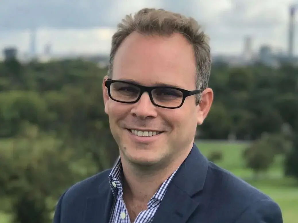 Nick Sutton of Sky News wearing suit and glasses smiling at the camera in front of a backdrop of a field and trees