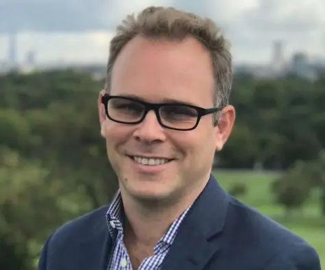 Nick Sutton of Sky News wearing suit and glasses smiling at the camera in front of a backdrop of a field and trees