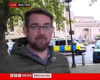 Screenshot from report in which BBC journalist Phillip Norton explained a protest was billed as being "pro-British". The ticker says "live - Bolton" and Norton is looking the camera wearing a lapel mic on his coat with police vans parked behind him