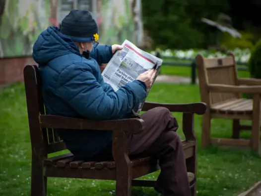 Man reading a local newspaper on a bench dressed warm in a coat and hat