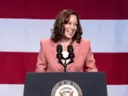 US Vice President Kamala Harris is seen stood laughing, looking off-camera in front of a large American flag and behind a podium.