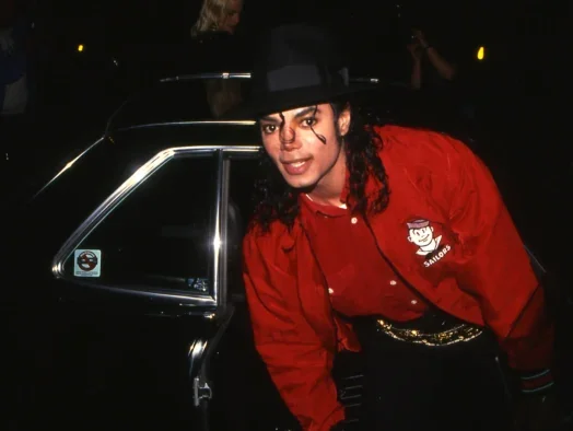 Michael Jackson bent over after getting out of a black car, behind him, and leaning towards the camera. He's wearing a black hat, with long hair down, and a red jacket