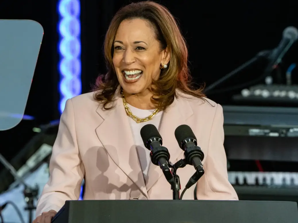 Vice President Kamala Harris standing at a lectern at night and laughing at something to the side of the camera. She's wearing a pale pink blazer, white round-necked top and gold necklace