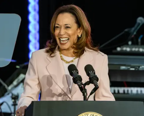 Vice President Kamala Harris standing at a lectern at night and laughing at something to the side of the camera. She's wearing a pale pink blazer, white round-necked top and gold necklace