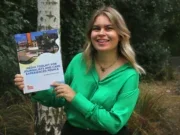 Sophia Alexandra Hall holds up Media Toolkit for Journalists and Care Experienced People book. She is smiling at the camera, wearing a green shirt, with a tree, grass and hedge in the background