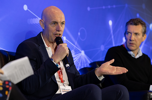 David Dinsmore speaking on stage holding a microphone and gesturing with his other hand. Wearing a suit jacket, white shirt and lanyard