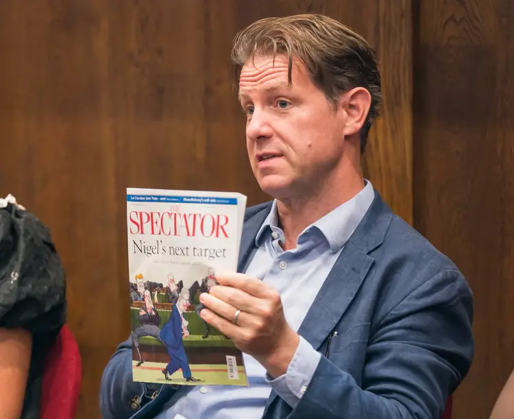 Fraser Nelson sits in a chair holding up a copy of The Spectator magazine. He's wearing a blazer and a shirt with no tie and appears to be mid-sentence