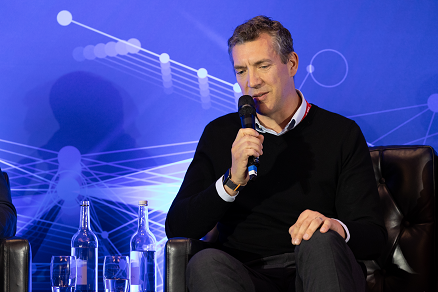 Graham Page sitting on a stage with a blue backdrop. He's holding a microphone and speaking, wearing a black jumper and trousers.