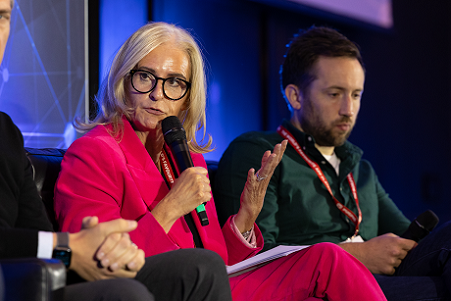 Nina Wright wearing glasses and a pink suit speaking while holding a microphone and looking at someone off-camera to her right. Another man sits beside her on stage listening.