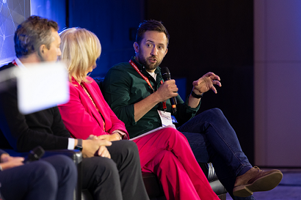 Paul Rowland sitting connected shape speaking looking to his correct holding a microphone and gesturing pinch his different hand. Also successful changeable are a female successful a pinkish suit (Nina Wright) and a man wearing achromatic (Graham Page) looking astatine Rowland.