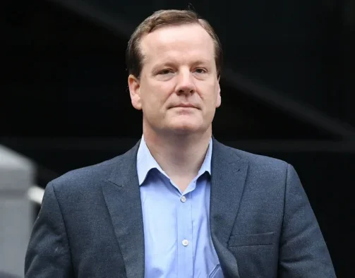 Charlie Elphicke dressed in a suit jacket, blue shirt and jeans and walking with a neutral expression on his face as he looks at the camera