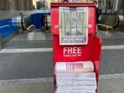 Picture shows red newspaper bin containing a stack of newspapers and a pinned up copy of the Evening Standard at the top to show its front page. Bin is in front of a backdrop of escalators leading down into a tube station.