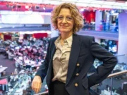 Naja Nielsen stands on a balcony in front of the BBC newsroom with rows of desks behind her. She's leaning on the balcony railing and looking at the camera, wearing a suit jacket and blouse