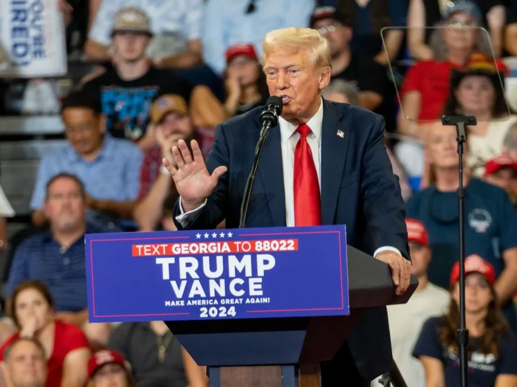 Donald Trump at a rally in August 2024 behind a podium with Trump/Vance sign and people sitting on bleachers behind him. He's talking into the mic and gesturing with one hand