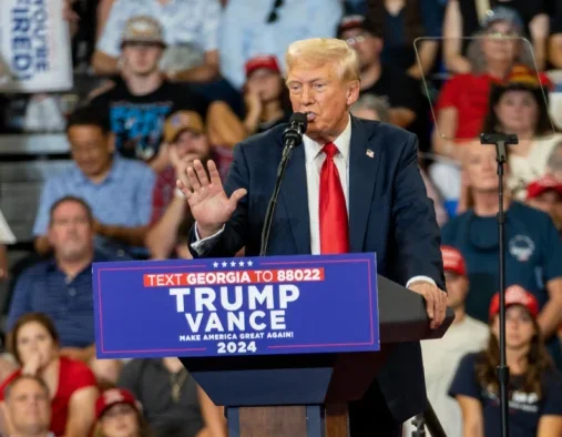 Donald Trump at a rally in August 2024 behind a podium with Trump/Vance sign and people sitting on bleachers behind him. He's talking into the mic and gesturing with one hand