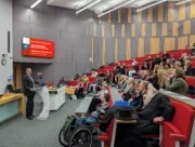 Bloomberg editor-in-chief John Micklethwait appears in front of a crows in a lecture hall at City University, London, where he speaks about journalism business models, paywalls, the effects of AI and lawfare. Also visible, in the third row from the front, is his wife, actress Kristin Scott Thomas.