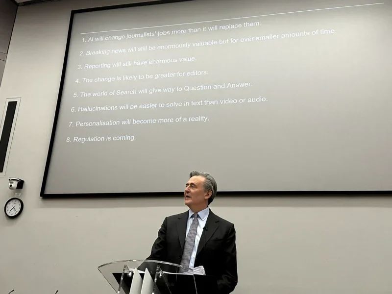 Bloomberg editor-in-chief John Micklethwait appears in front of a projector screen showing his eight predictions for journalism and AI.