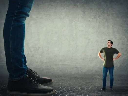 Small man stands with hands on hips looking up at a giant, of which you can only see his massive legs and shoes.