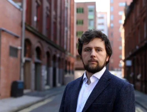 Sam McBride standing in a street with red-brick buildings behind, wearing a jacket and white shirt