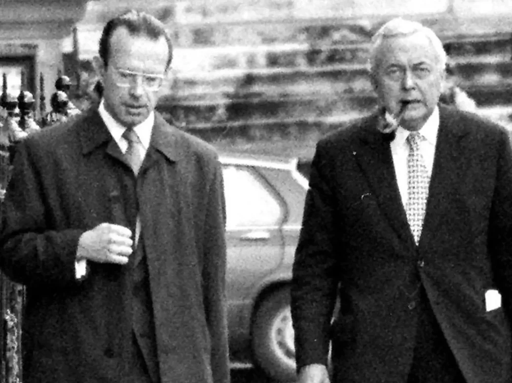 Black and white photo of Joe Haines dressed in a long coat and a suit underneath with big glasses, walking next to Harold Wilson with white hair, a pipe in his mouth, and wearing a suit. They are walking down a London street side by side.