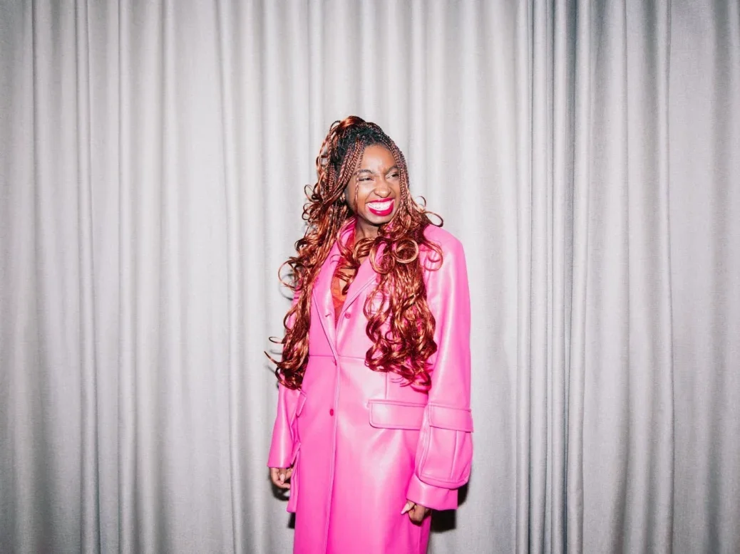 Kemi Alemoru, the new head of editorial content at Glamour UK, stands grinning and looking off-camera in front of a grey curtain and wearing a hot pink trenchcoat.