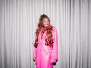 Kemi Alemoru, the new head of editorial content at Glamour UK, stands grinning and looking off-camera in front of a grey curtain and wearing a hot pink trenchcoat.