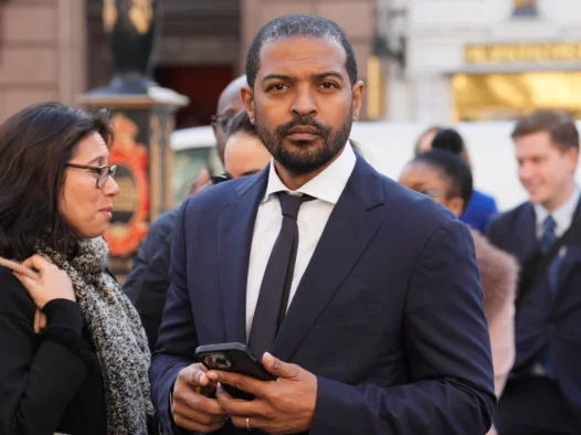 Noel Clarke dressed in a suit holding a phone standing in front of people behind him on the street and staring into the camera