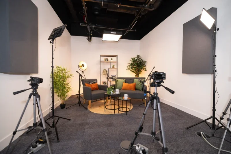 Video studio set up in 'cosy' way with armchair, sofa, two large plants and homely shelving unit behind. Also two cameras on tripods, various lighting rigs.