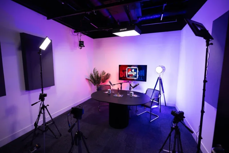 Video/podcast studio with TV backdrop featuring The Division Bell podcast branding. Table and two chairs plus cameras and lighting all present.