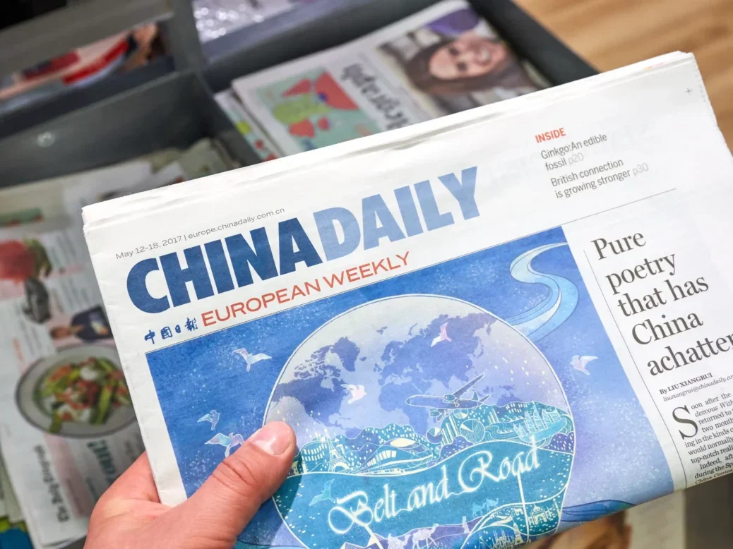 A copy of China Daily from 2017 is held up to the camera above a stack of newspapers. The front page carries an illustration promoting China's Belt and Road Initiative, and the top headline reads: "Pure poetry that has China achatter". It is accompanied by smaller headlines including "Ginkgo: an edible fossil" and "British connection is growing stronger".