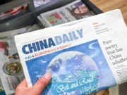 A copy of China Daily from 2017 is held up to the camera above a stack of newspapers. The front page carries an illustration promoting China's Belt and Road Initiative, and the top headline reads: "Pure poetry that has China achatter". It is accompanied by smaller headlines including "Ginkgo: an edible fossil" and "British connection is growing stronger".