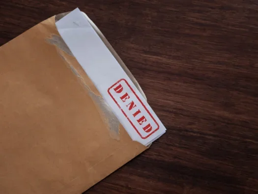 A manila envelope is seen torn open on a wooden table with a white document peeking out stamped with the word: "DENIED" in red. The picture illustrates a story about Sheffield academic Jingrong Tong's latest study, which concludes based on interviews with 31 UK journalists about the state of Freedom of Information that the UK is at risk of becoming a propaganda state over time.