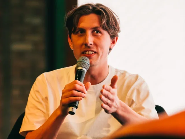 Mundial editor James Bird wearing white t-shirt, holding a microphone and gesturing with the other hand while talking